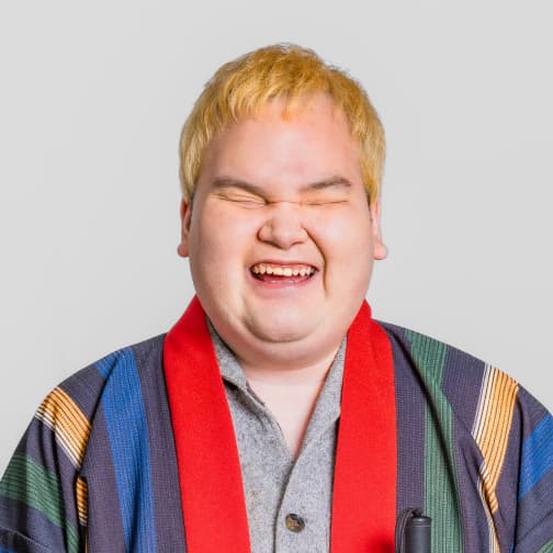 北村直也のポートレート。金色の短髪で、目を閉じて笑っている。緑、黄、青、濃紺の縞模様と赤い襟の法被を着ている。