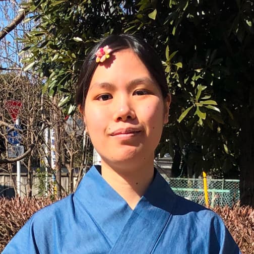 関場理生のポートレート。淡い藍色の着物を着て、花の髪飾りをつけている。木々を背に屋外で正面を向いている。